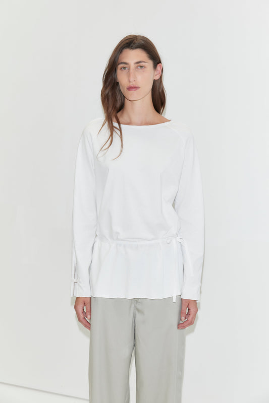 A woman with long dark hair stands against a plain white background wearing the Wided Longsleeve - White by Deiji Studios, a white top with a wide neckline and drawstring hem, with grey wide-leg trousers.