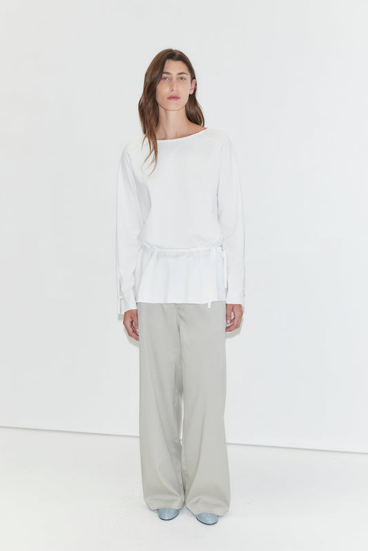 A woman with long dark hair stands against a plain white background wearing the Wided Longsleeve - White by Deiji Studios, a white top with a wide neckline and drawstring hem, with grey wide-leg trousers and pale blue heels.
