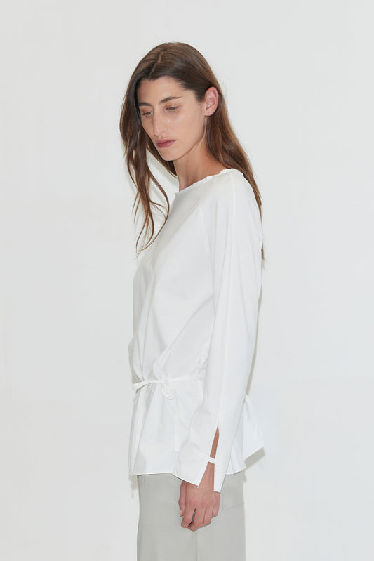 A woman with long dark hair stands in profile against a plain white background, looking down, wearing the Wided Longsleeve - White by Deiji Studios, a white top with a wide neckline and drawstring hem, with grey trousers.