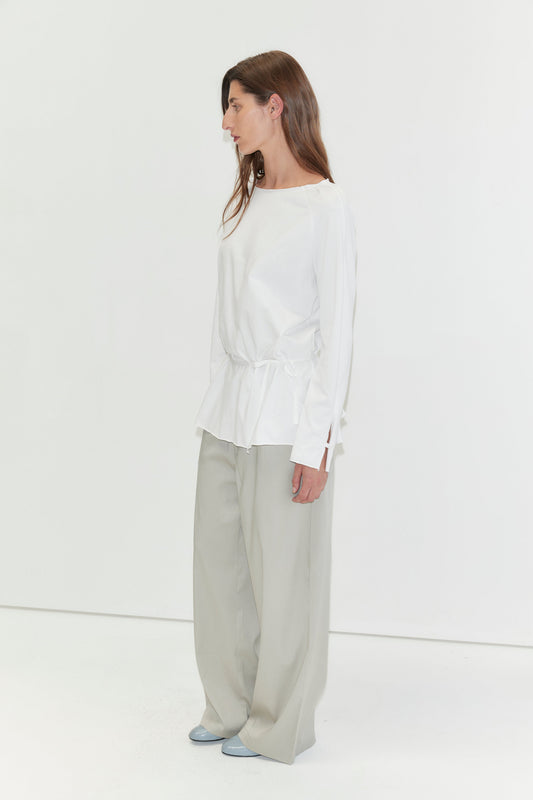 A woman with long dark hair stands in profile against a plain white background wearing the Wided Longsleeve - White by Deiji Studios, a white top with a wide neckline and drawstring hem, with grey wide-leg trousers and pale blue heels.