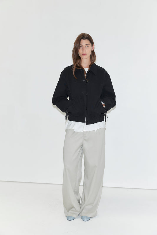 A person stands against a plain white background, wearing a black jacket over a white shirt, the Tencel Pant - Grey from Deiji Studios styled as loose wide-leg pants, and light blue shoes, with their hands in their jacket pockets.