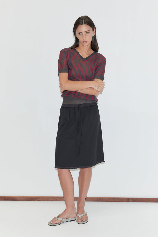 A woman with long dark hair stands against a plain white background with arms crossed, wearing the One Frill Skirt - Black by Deiji Studios, a black knee-length skirt with a drawcord waist and single frill hem, with a red striped knit tee and flip-flops.