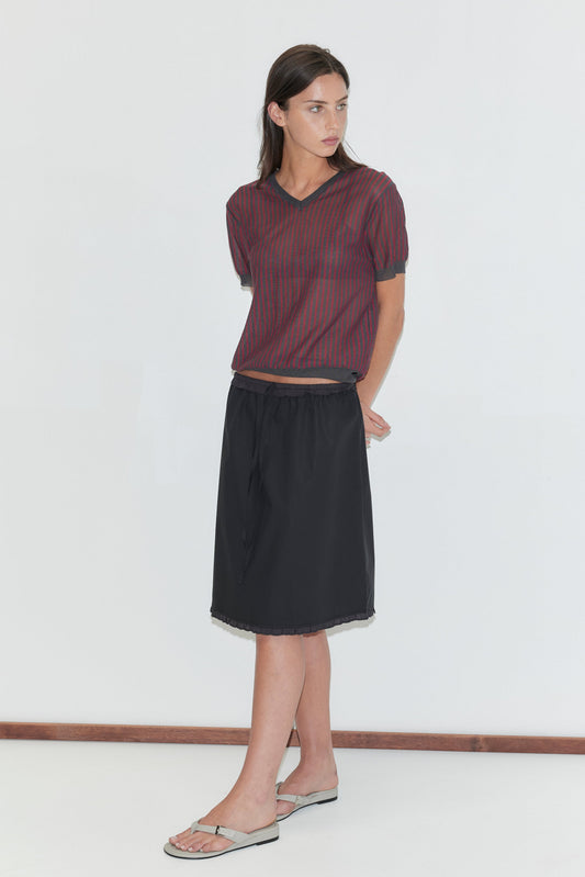 A woman with long dark hair stands against a plain white background wearing the One Frill Skirt - Black by Deiji Studios, a black knee-length skirt with a drawcord waist and single frill hem, with a red striped knit tee and flip-flops.