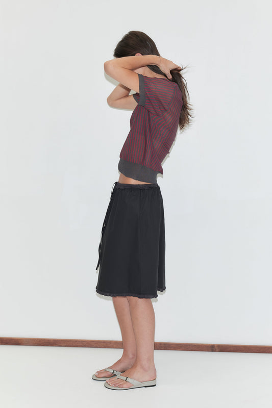 A woman with long dark hair stands in profile against a plain white background wearing the One Frill Skirt - Black by Deiji Studios, a black knee-length skirt with a drawcord waist and frill hem, with a red striped knit tee and flip-flops.
