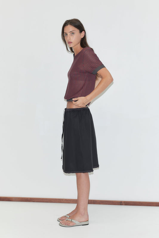 A woman with long dark hair stands in profile against a plain white background wearing the Cross Neck Tee - Red Stripe by Deiji Studios, a fine red and charcoal striped knit tee, with a black midi skirt and grey flip-flops.