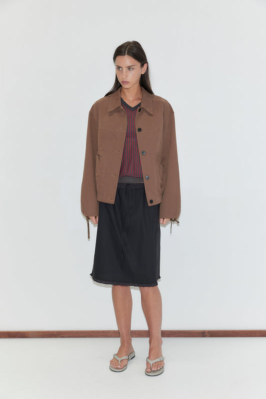 A woman with long dark hair stands against a plain white background wearing the One Frill Skirt - Black by Deiji Studios, a black knee-length skirt with a frill hem, with a red striped knit tee, a brown jacket and flip-flops.