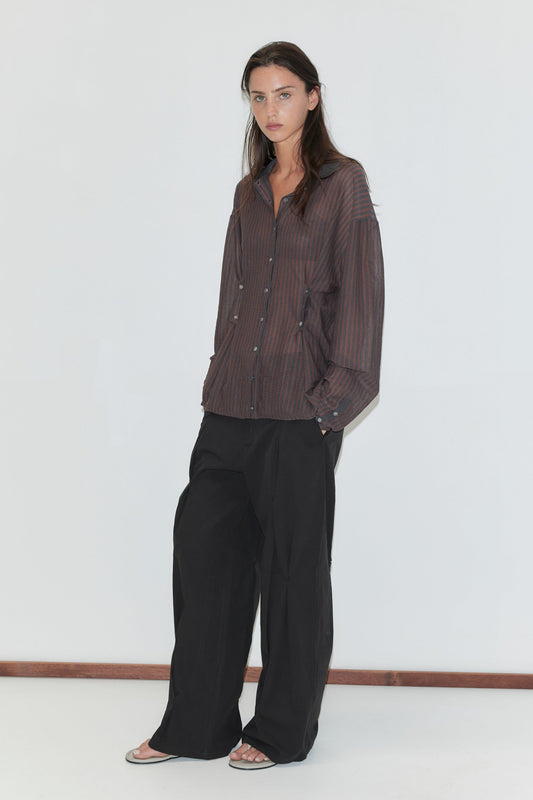 A woman with long dark hair stands against a plain white background wearing the Multi Pleat Pant - Black by Deiji Studios, black wide-leg pleated trousers, with a sheer brown striped knit shirt and flip-flops.