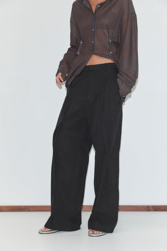 A cropped view of the Multi Pleat Pant - Black by Deiji Studios, black wide-leg trousers with front pleats and a waistband, worn with a sheer brown striped knit shirt against a plain white background.
