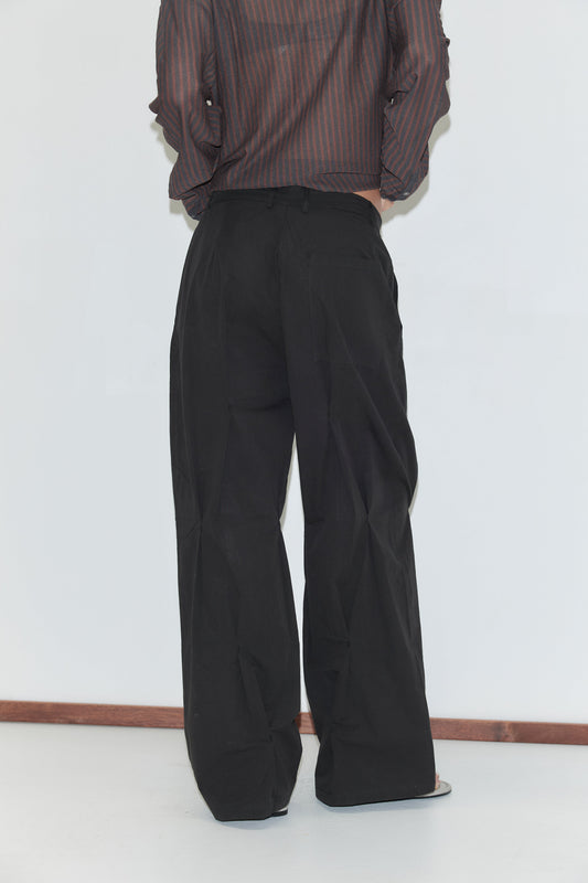 A cropped back view of the Multi Pleat Pant - Black by Deiji Studios, black wide-leg pleated trousers with back welt pockets, worn with a sheer brown striped knit shirt against a plain white background.