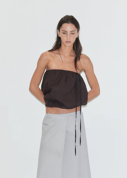 A woman with long dark hair and a neutral expression stands against a plain white background with her hands behind her back, wearing the Bubble Top - Brown Black Stripe by Deiji Studios, a strapless bubble-hem linen top with a contrast black drawcord tie, paired with a light grey striped wrap skirt.