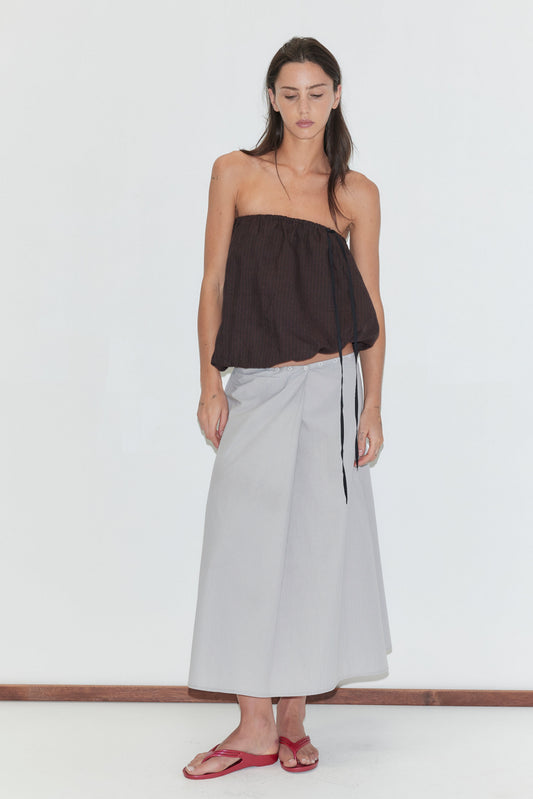 A woman with long dark hair stands against a plain white background wearing the Wrap Skirt - Cloud Stripe by Deiji Studios, a pale grey-blue fine-striped wrap midi skirt, with a brown strapless bubble top and red flip-flops.