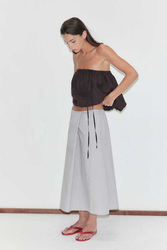 A woman with long dark hair stands against a plain white background looking down, wearing the Wrap Skirt - Cloud Stripe by Deiji Studios, a pale grey-blue fine-striped wrap midi skirt, with a brown strapless bubble top and red flip-flops.
