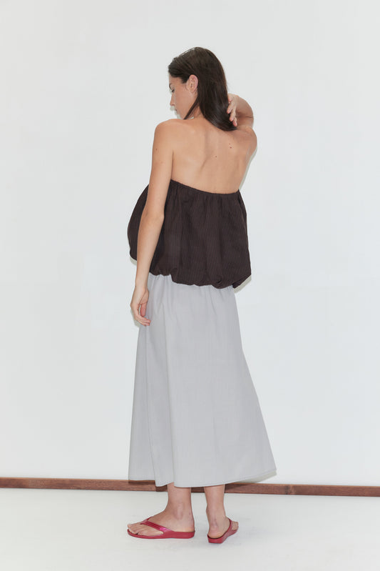 A woman with long dark hair stands with her back to the camera, wearing the Bubble Top - Brown Black Stripe by Deiji Studios and a light grey striped wrap skirt with red flip-flops, against a plain white background.