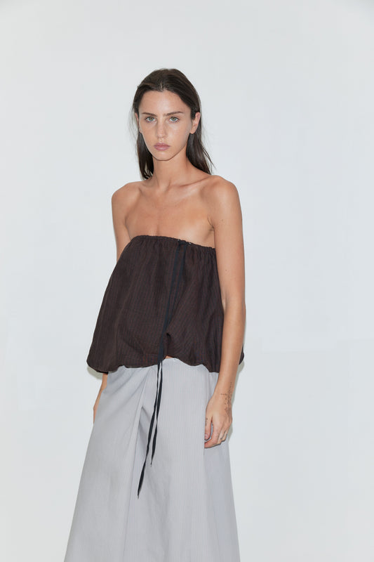 A cropped view of a woman wearing the Wrap Skirt - Cloud Stripe by Deiji Studios, a pale grey-blue fine-striped wrap skirt with a front overlap, and a brown strapless bubble top, against a plain white background.