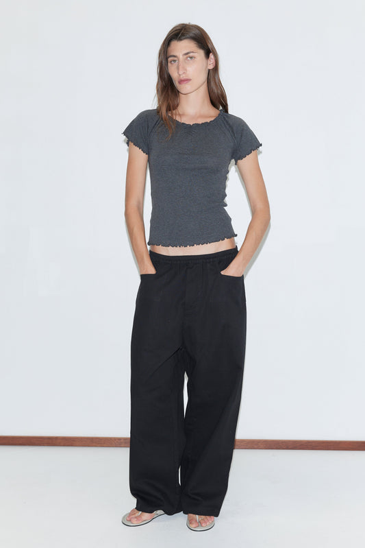 A woman with long brown hair and a neutral expression stands against a white background, wearing the Square Pocket Pant in black—relaxed organic cotton twill pants with square pockets—paired with a fitted dark gray short-sleeved top and flat sandals.