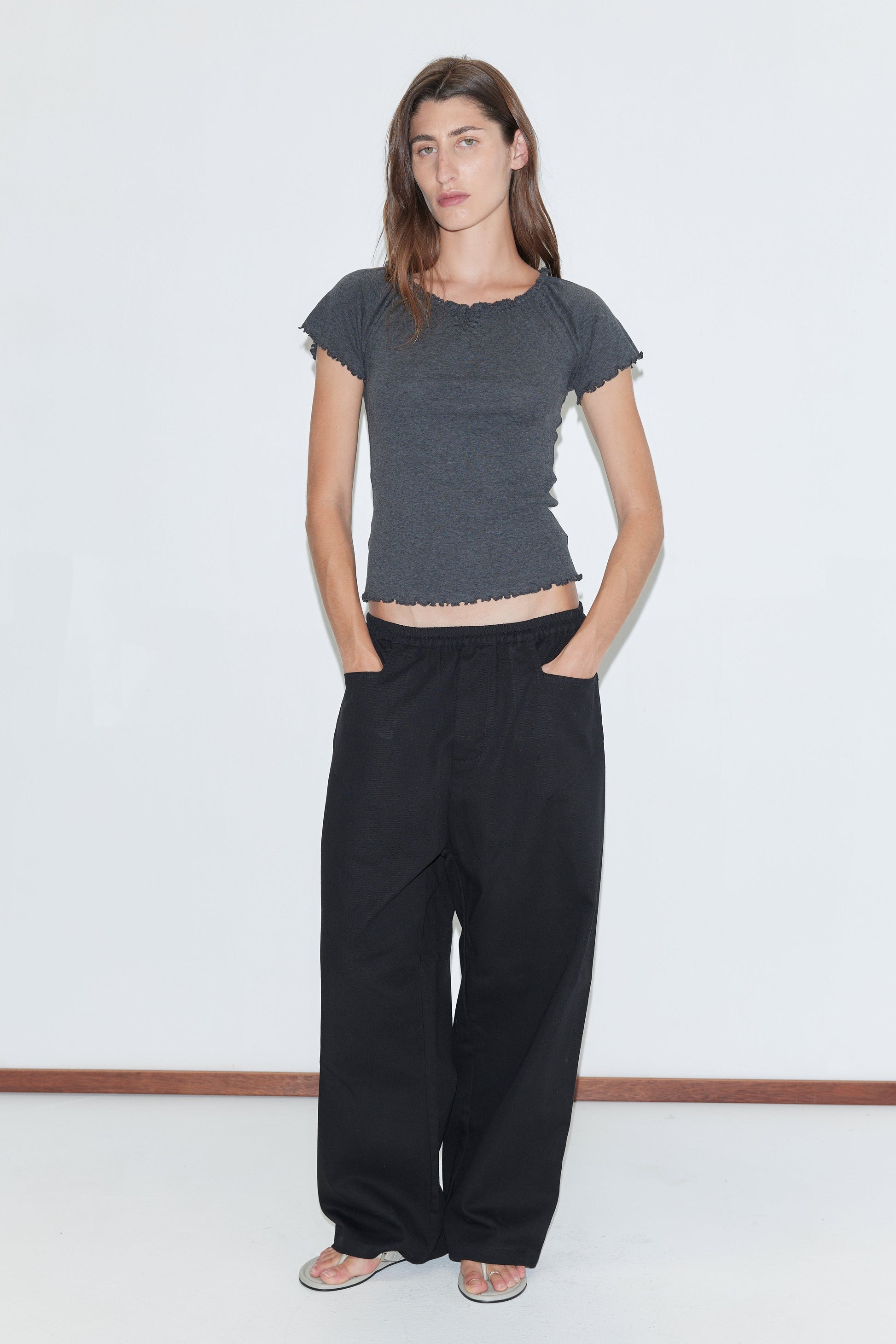 A woman with long brown hair and a neutral expression stands against a white background, wearing the Square Pocket Pant in black—relaxed organic cotton twill pants with square pockets—paired with a fitted dark gray short-sleeved top and flat sandals.