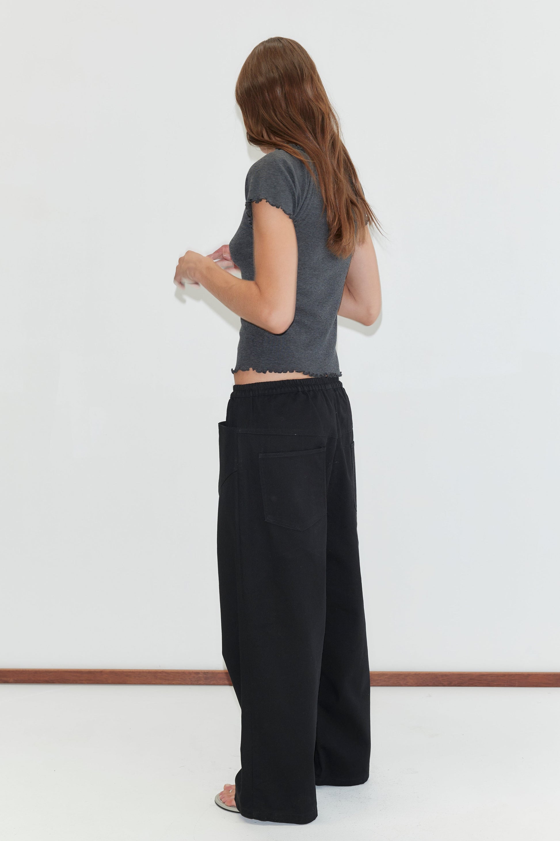 A woman with long brown hair, in a gray short-sleeve shirt and Square Pocket Pant - Black, stands barefoot facing away against a plain white wall.
