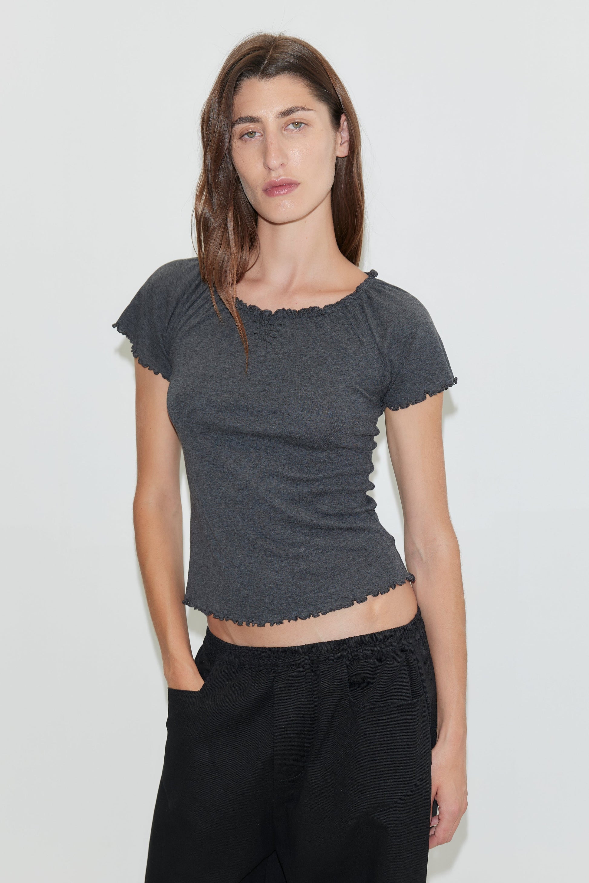 A woman with long brown hair wears the Square Pocket Pant - Black, styled with a fitted dark gray short-sleeve top. She stands against a plain white wall, one hand in her pocket, looking directly at the camera.