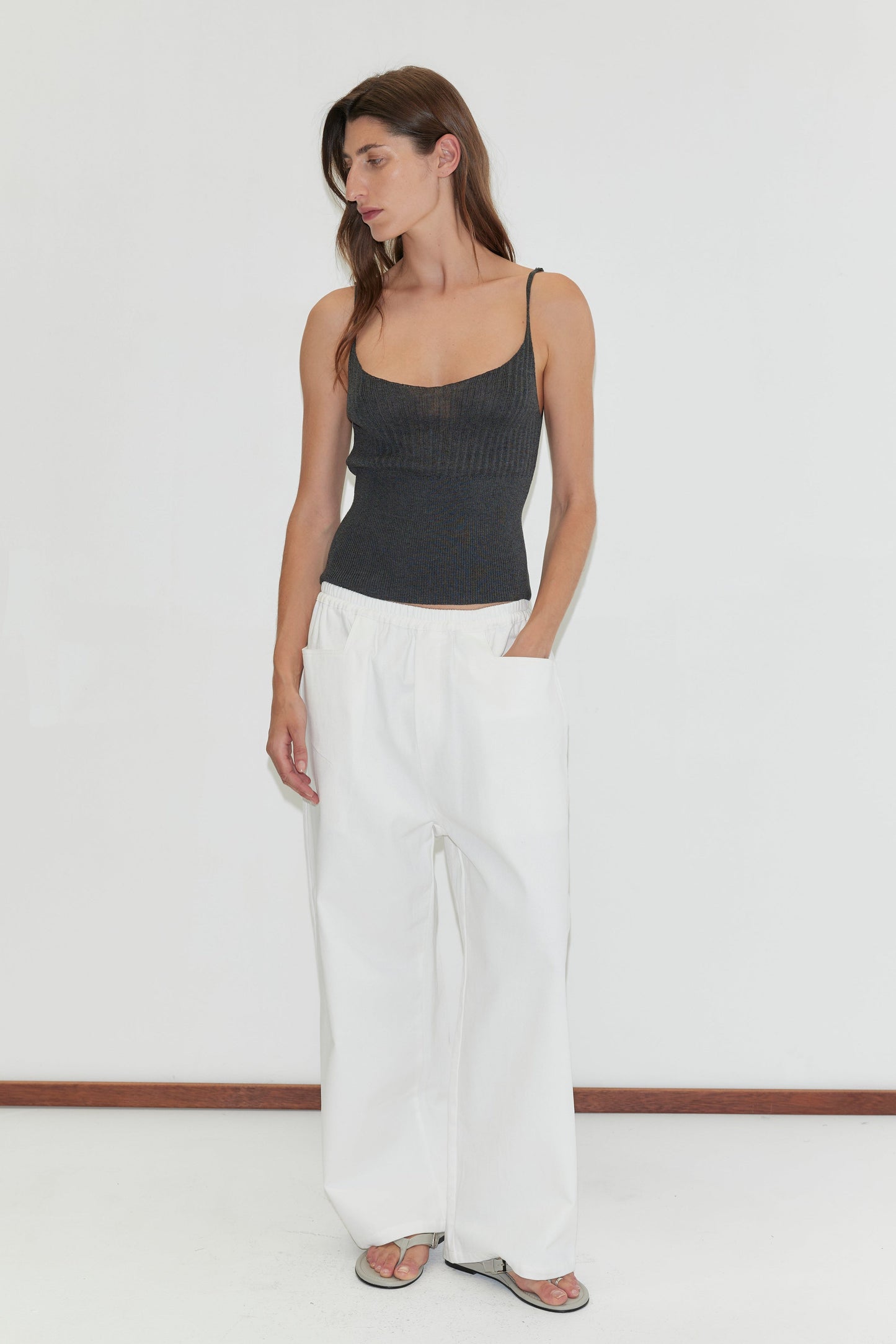 A woman with straight brown hair stands against a white background, looking down to her left with one hand in the pocket of her Square Pocket Pant in white. She pairs the relaxed, straight-leg pant with a dark gray tank top and sandals.