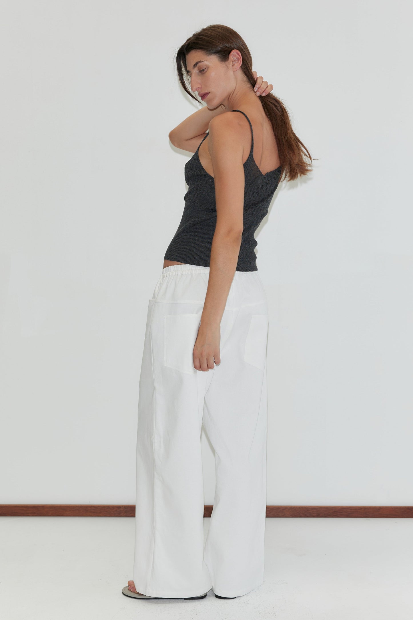 A woman with long brown hair stands sideways against a white background, wearing the Square Pocket Pant in White with a straight leg silhouette and a dark sleeveless top. She touches the back of her neck while looking down.
