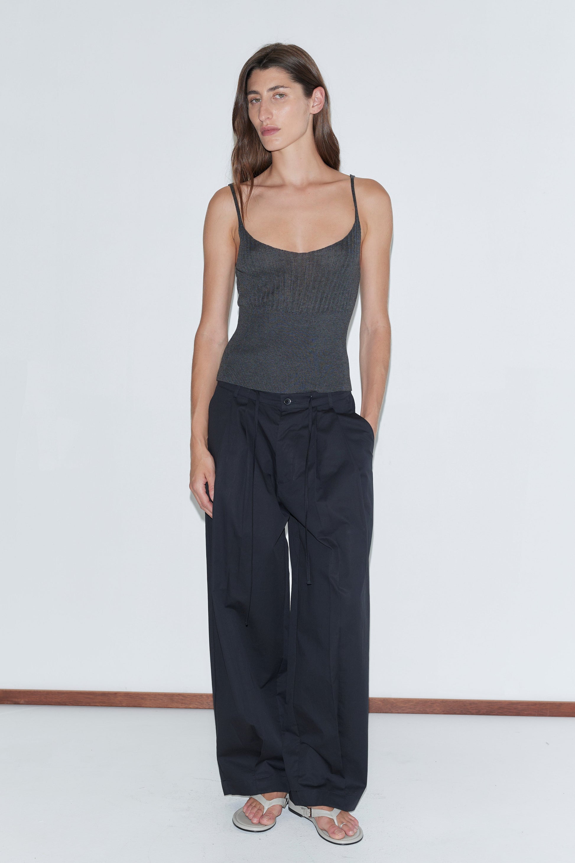 A person stands against a white wall, wearing a Deiji Studios dark gray tank top and the Turn Pant - Black with an adjustable drawcord, hands in pockets, sandals on, long brown hair flowing and a neutral expression.