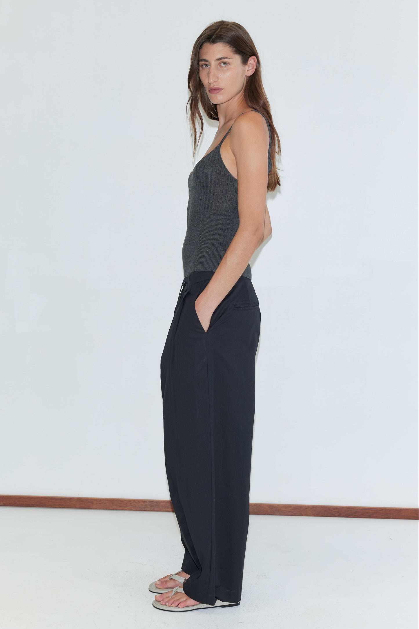 A person with long brown hair, wearing a dark gray sleeveless top, Turn Pant - Black by Deiji Studios with an adjustable drawcord, and flip-flops, stands sideways with hands in pockets against a plain white background.