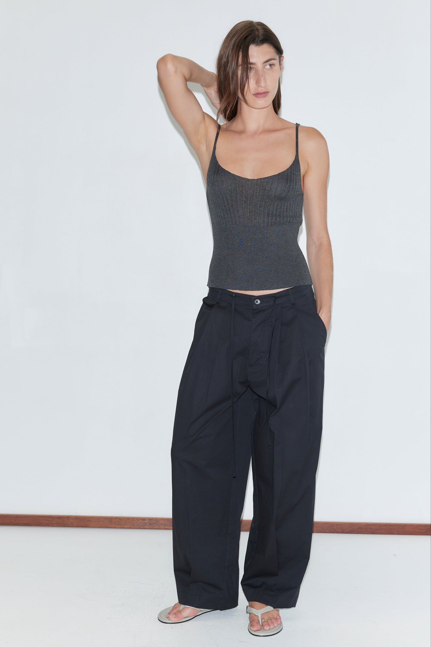 A woman stands against a plain white wall, one hand behind her head, wearing a dark gray tank top, Deiji Studios Turn Pant in black with an adjustable drawcord, and sandals. Her expression is neutral, hair parted to the side.