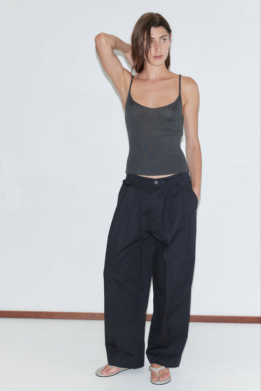 A woman stands against a plain white wall, one hand behind her head, wearing a dark gray tank top, Deiji Studios Turn Pant in black with an adjustable drawcord, and sandals. Her expression is neutral, hair parted to the side.