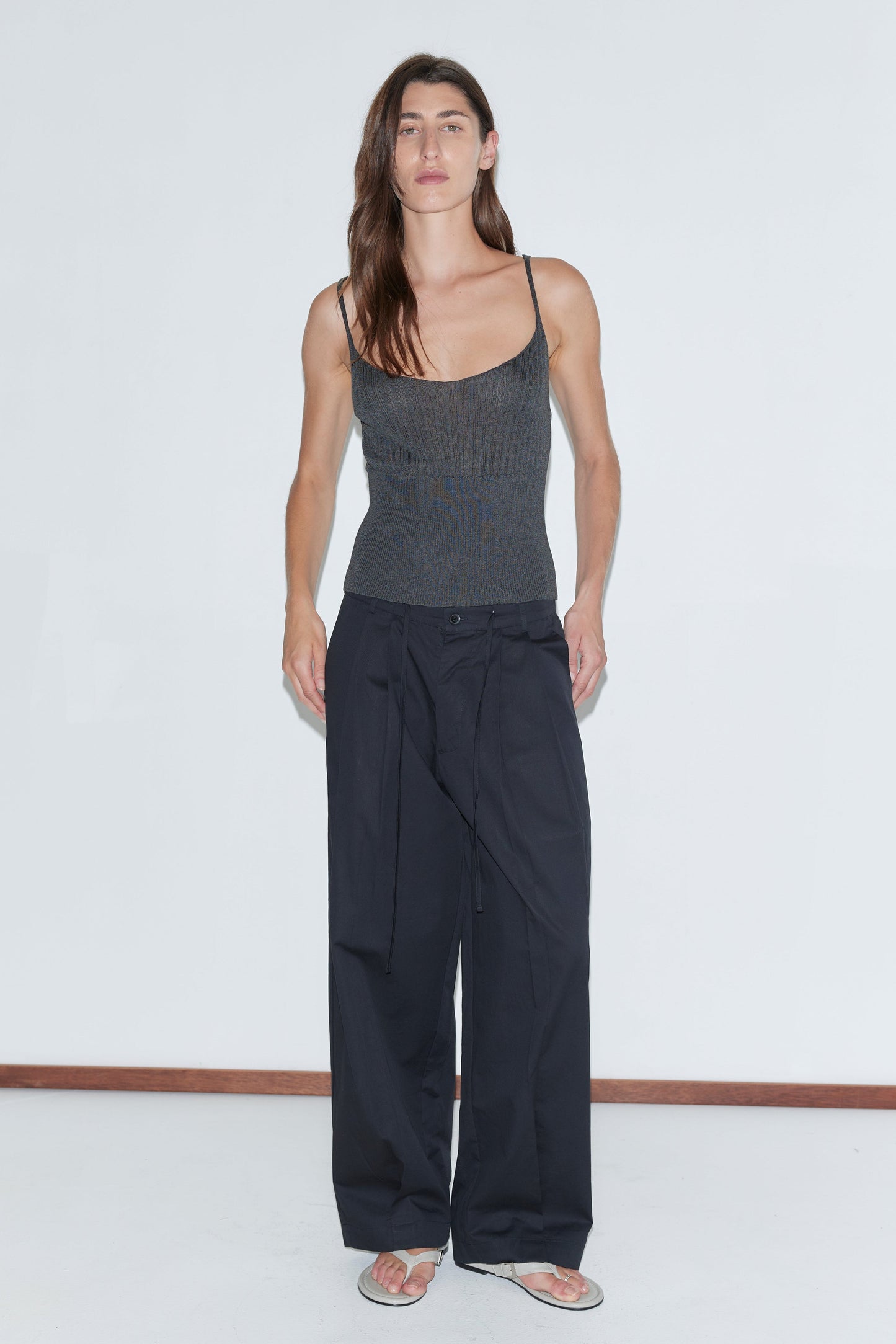 A person with long brown hair wears a fitted dark spaghetti-strap top, the Turn Pant - Black by Deiji Studios with an adjustable drawcord waist, and beige flip-flops, standing against a plain white background.