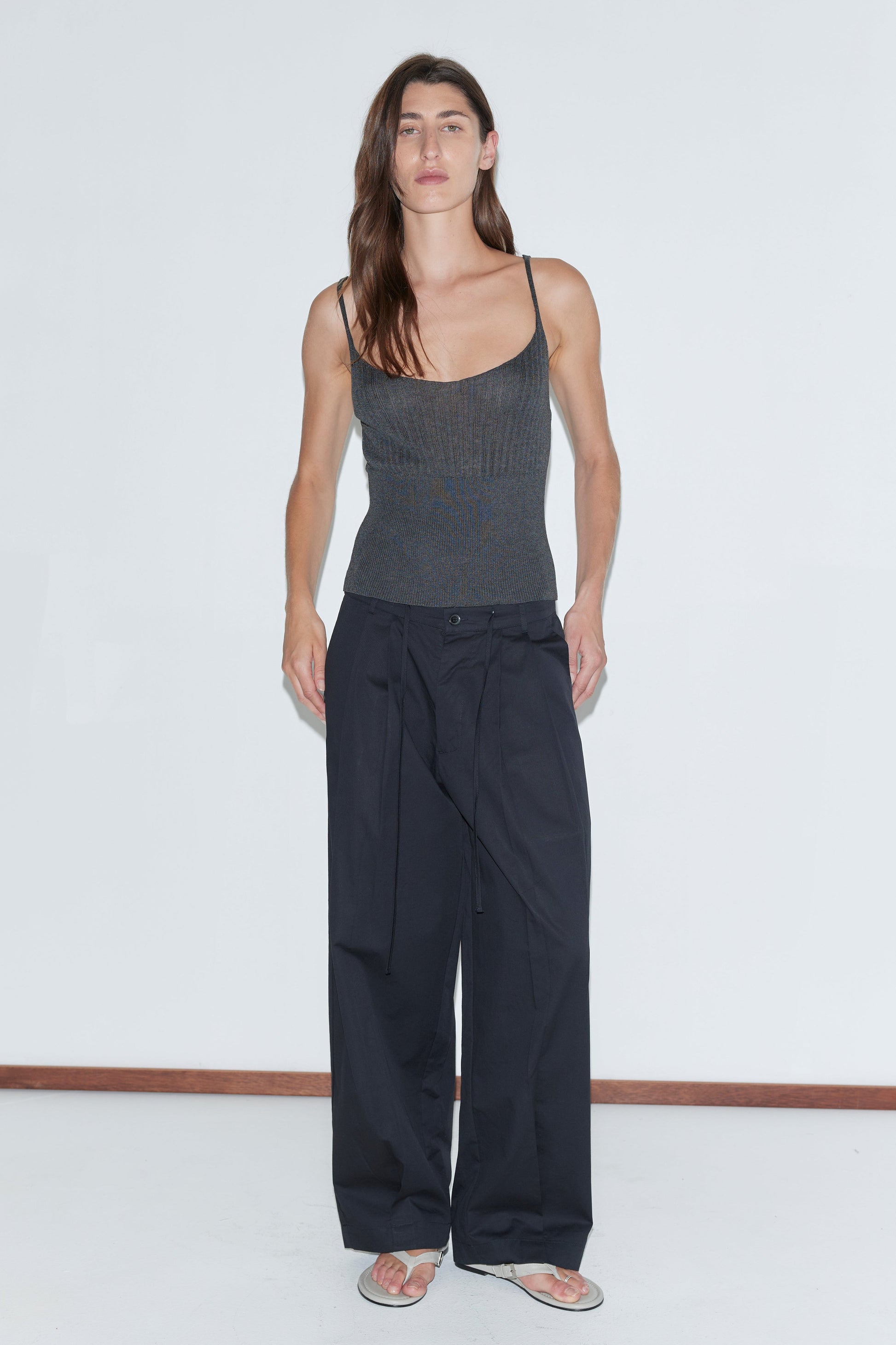 A person with long brown hair wears a fitted dark spaghetti-strap top, the Turn Pant - Black by Deiji Studios with an adjustable drawcord waist, and beige flip-flops, standing against a plain white background.