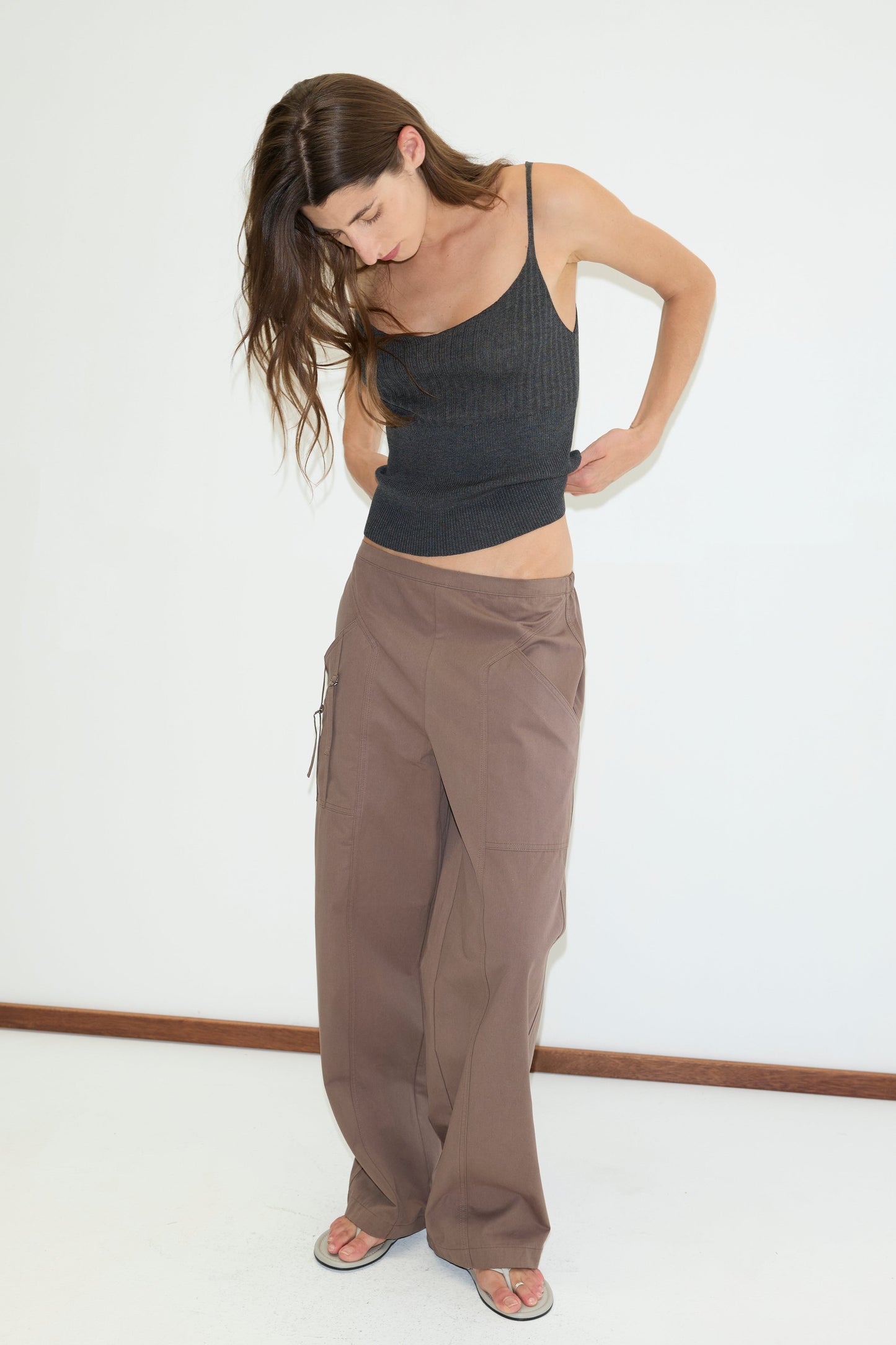 A woman with long brown hair wears a fitted black tank top, the Adjust Pocket Pant in Sawdust, and sandals while looking down and standing against a plain white wall.
