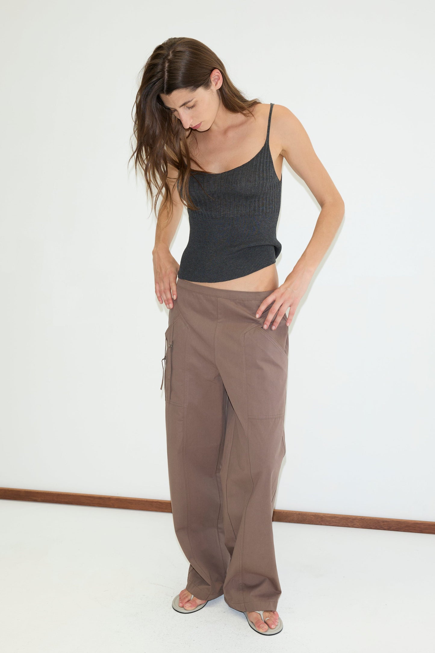 A woman with long brown hair, barefoot, wears a black tank top and Deiji Studios’ Adjust Pocket Pant in Sawdust as she adjusts the waistband. She stands on a white floor against a plain white wall.