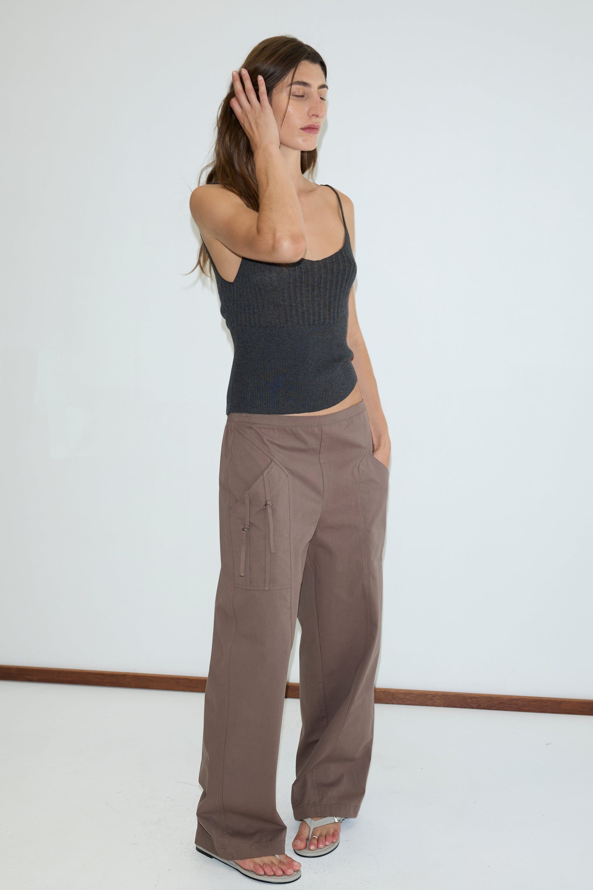 A woman with long brown hair stands by a white wall in a dark knit tank, Adjust Pocket Pant - Sawdust by Deiji Studios, and white flip-flops. She touches her face with one hand, looking relaxed.