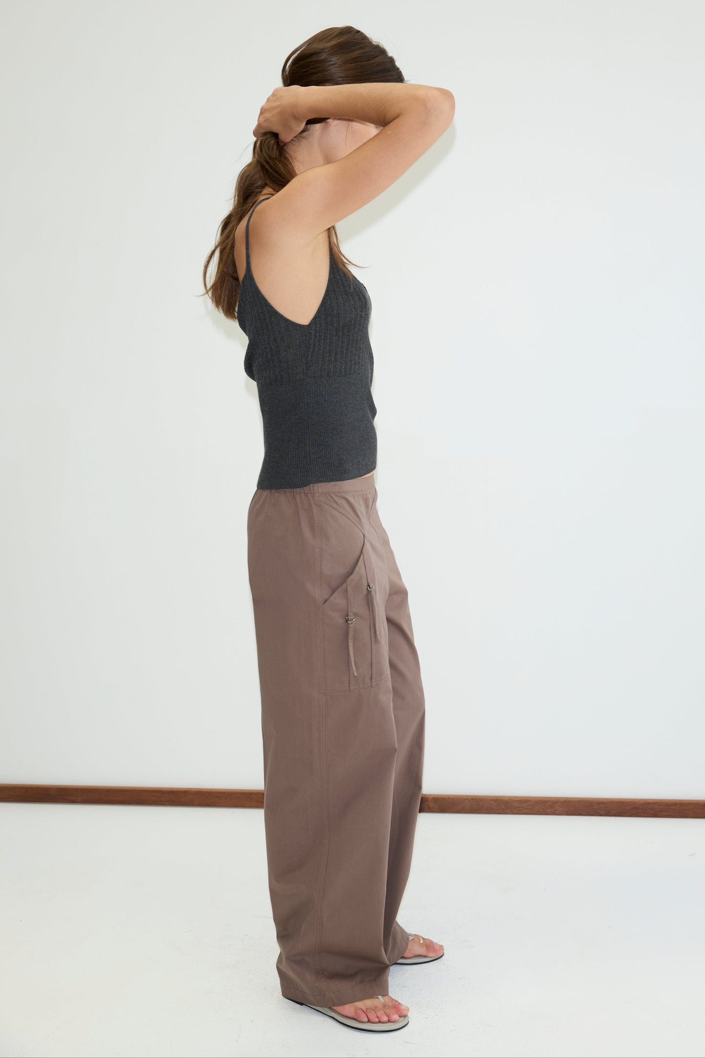 A woman stands sideways against a plain white wall, wearing a dark sleeveless top, Deiji Studios Adjust Pocket Pant in Sawdust, and sandals. She has one arm raised to her head and her long hair behind her shoulders.