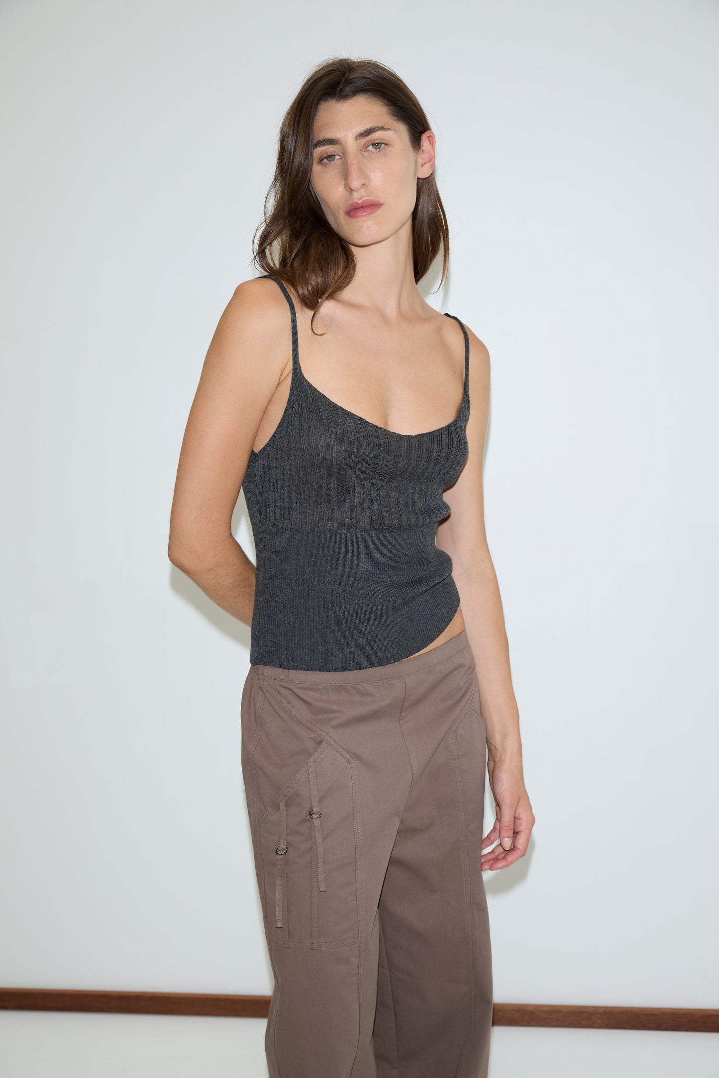 A woman with long brown hair stands against a plain white background, wearing a Deiji Studios dark gray sleeveless knit top and Adjust Pocket Pant - Sawdust. She looks at the camera with a neutral expression.