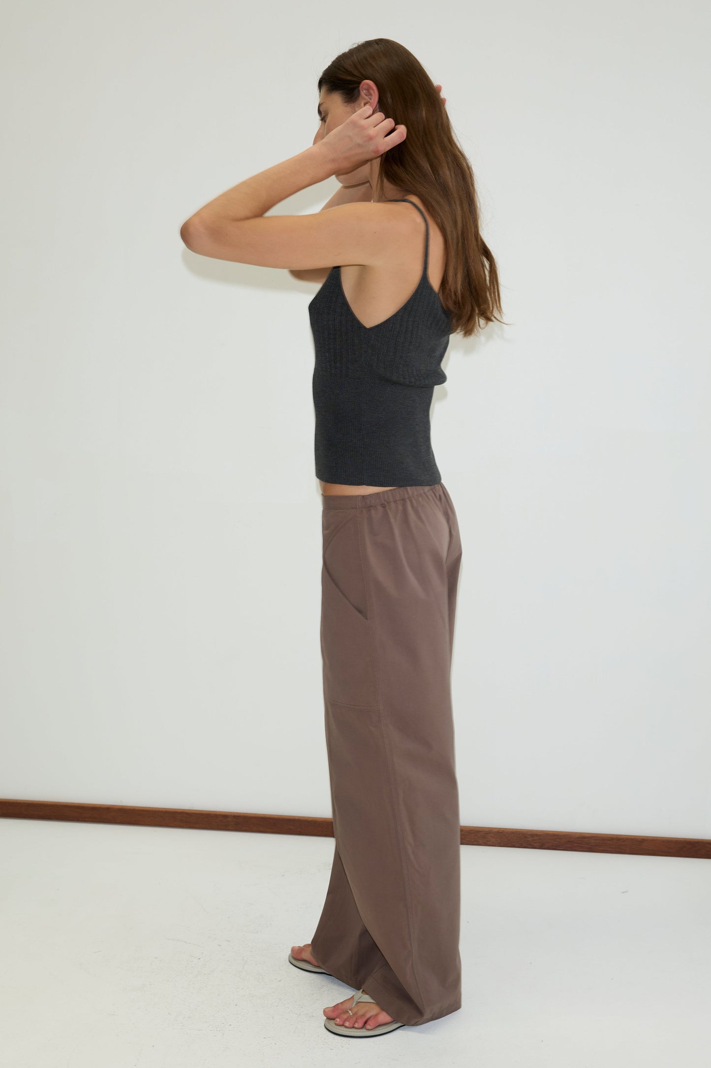 A woman with long brown hair stands sideways against a white wall, wearing a sleeveless dark gray top and Deiji Studios Adjust Pocket Pant - Sawdust with sandals, touching the side of her head with both hands.
