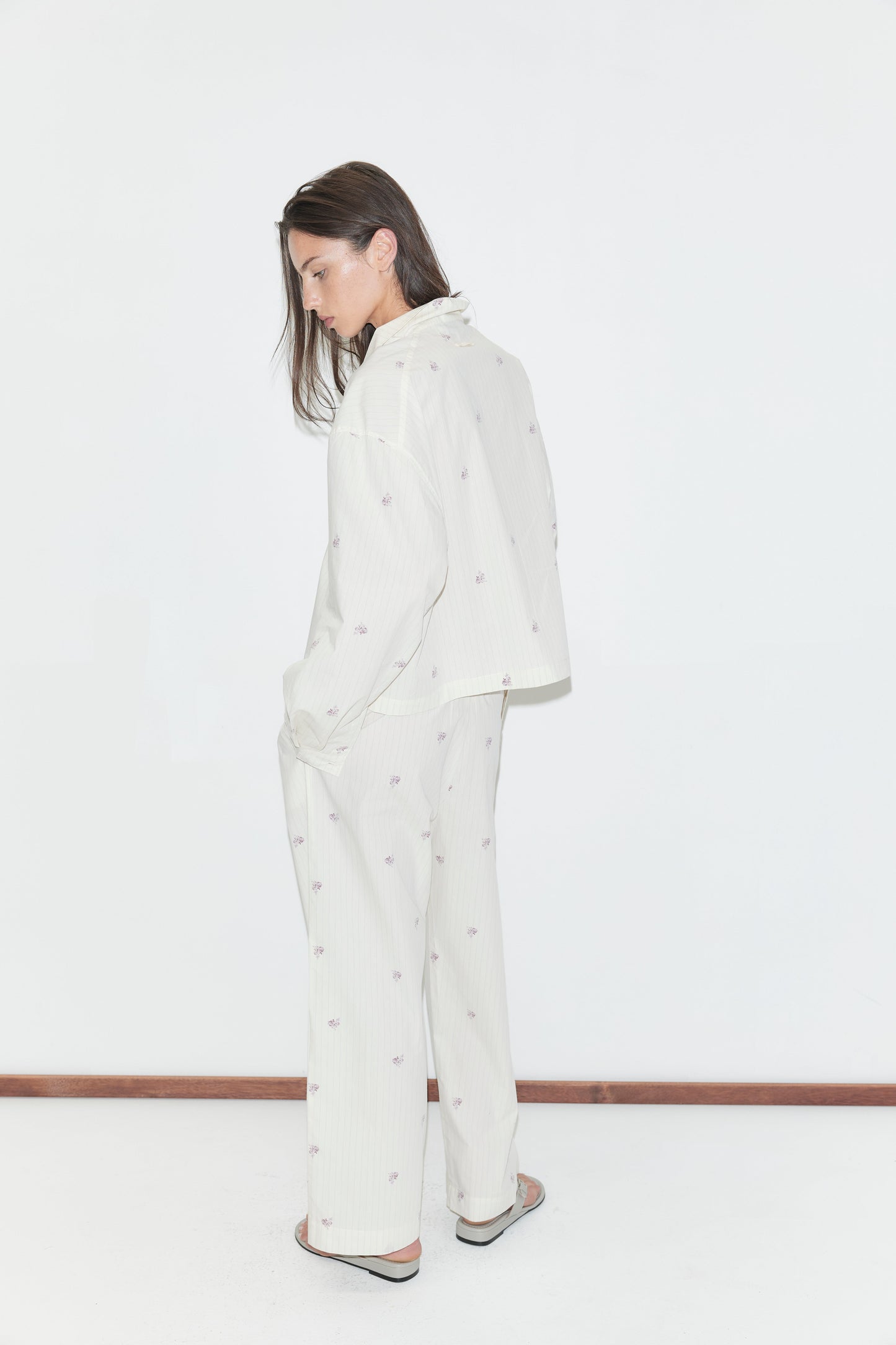 A person stands sideways against a white background, wearing the Ease Trouser - Floral Stripe pajama set, which features a loose fit with straight-leg trousers adorned with small, subtle floral stripe patterns. Their arms hang relaxed by their sides, and their long brown hair falls forward as they look down slightly.