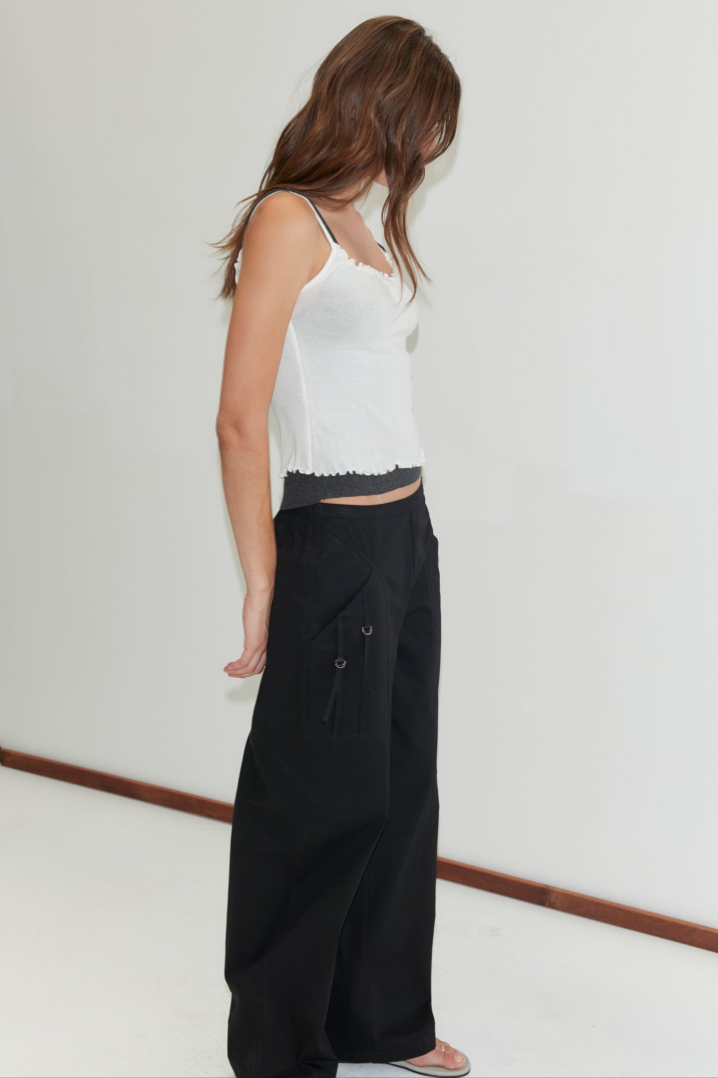 A woman with long brown hair stands against a white wall, wearing a Deiji Studios white tank top and Adjust Pocket Pant - Black. She looks down and to the side, arms relaxed by her sides.