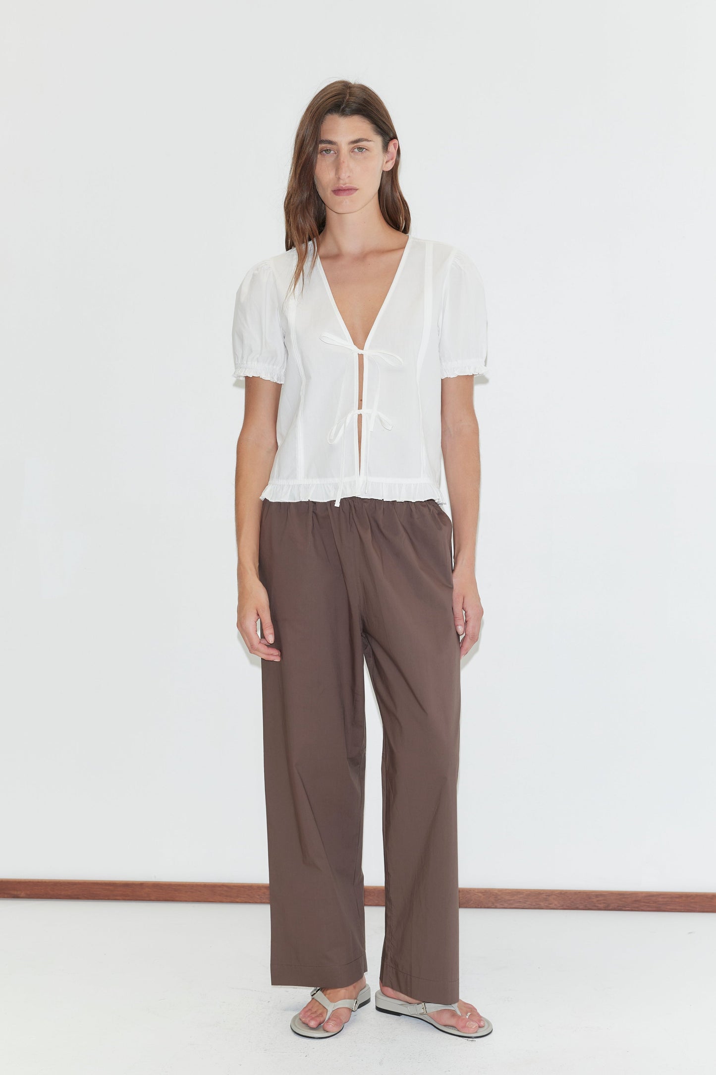 A person with long brown hair and a neutral expression stands against a white wall wearing a sheer white short-sleeve blouse, the Ease Trouser - Chocolate by Deiji Studios in organic cotton, and flat sandals.