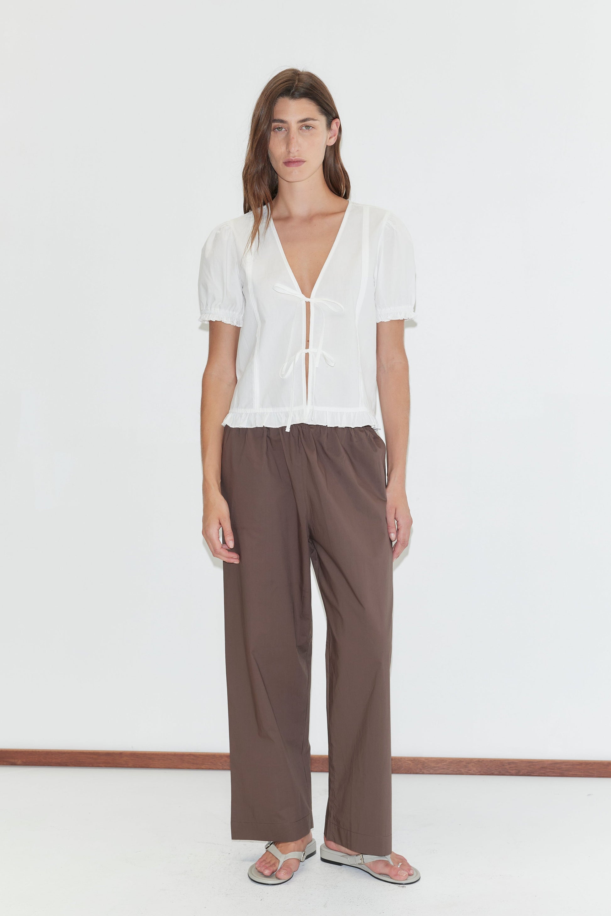 A person with long brown hair stands against a white background, wearing a white short-sleeve V-neck top with ruffled hem, Deiji Studios Ease Trouser - Chocolate, and sandals in a relaxed pose.