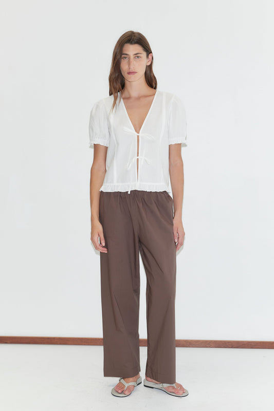 A person with long brown hair stands against a white background, wearing a white short-sleeve V-neck top with ruffled hem, Deiji Studios Ease Trouser - Chocolate, and sandals in a relaxed pose.
