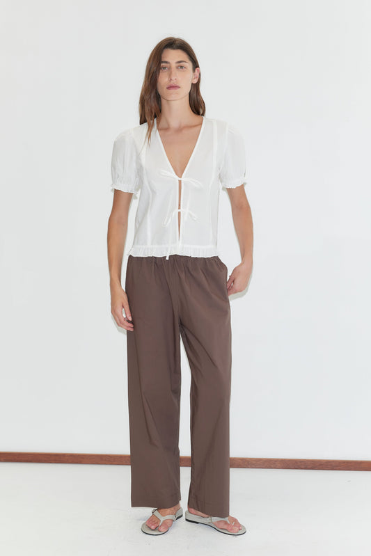 A person with long brown hair stands against a white wall, wearing a sheer blouse, the Ease Trouser - Chocolate by Deiji Studios in a wide-leg silhouette, and sandals.