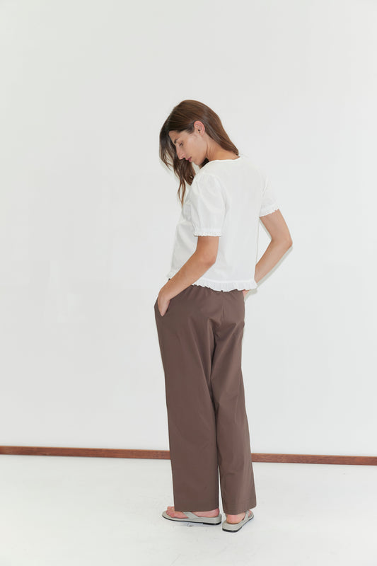A woman stands sideways, looking down, wearing the Pintuck Top - White by Deiji Studios with blouson sleeves, loose brown pants, and sandals against a plain white background.