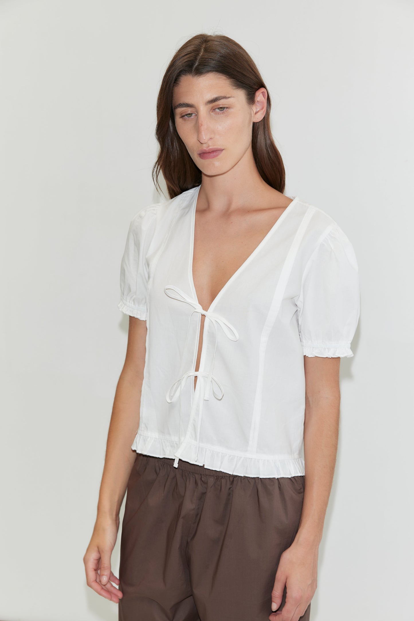 A woman with long brown hair wears the Deiji Studios Pintuck Top - White, featuring a deep V-neck and tie-front detail, paired with loose brown pants, standing against a plain white background.