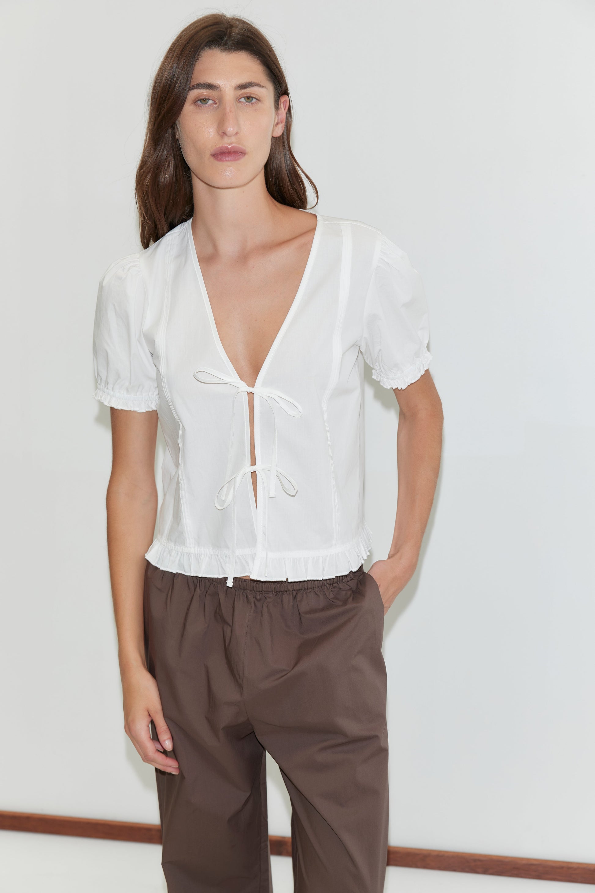 A person with long brown hair wears the Deiji Studios Pintuck Top in white with blouson sleeves and loose brown pants, standing against a plain white background with one hand in their pocket.