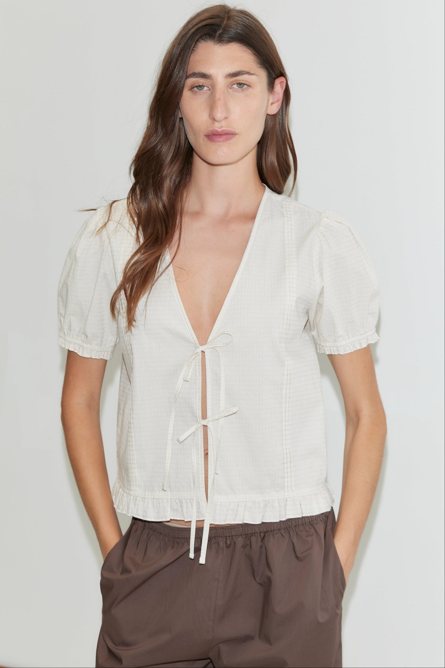 A woman with long brown hair stands against a white background, wearing the Pintuck Top - Bone Check by Deiji Studios with tie details and puffed sleeves, paired with loose brown pants. Her hands are in her pockets.