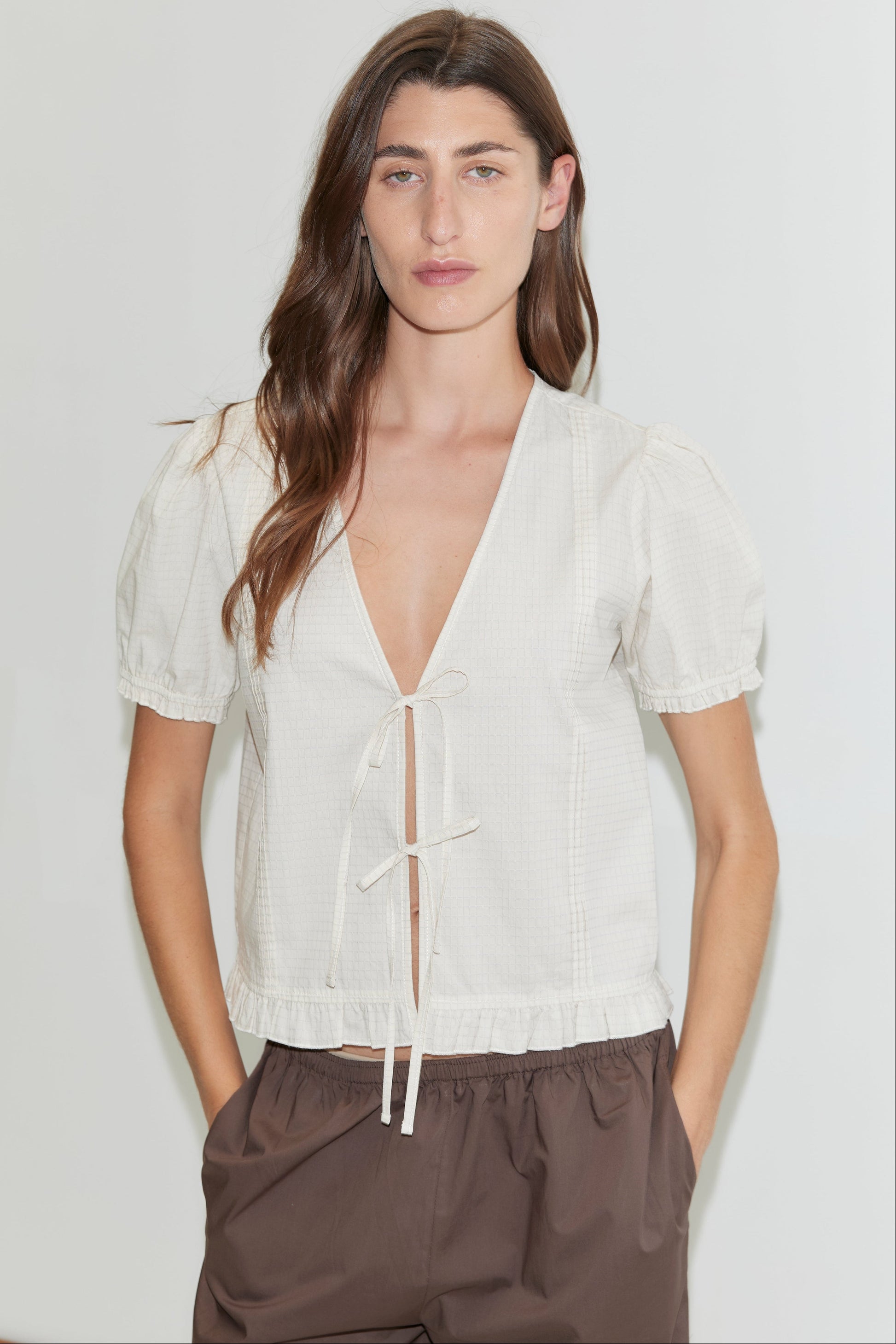 A woman with long brown hair stands against a white background, wearing the Pintuck Top - Bone Check by Deiji Studios with tie details and puffed sleeves, paired with loose brown pants. Her hands are in her pockets.