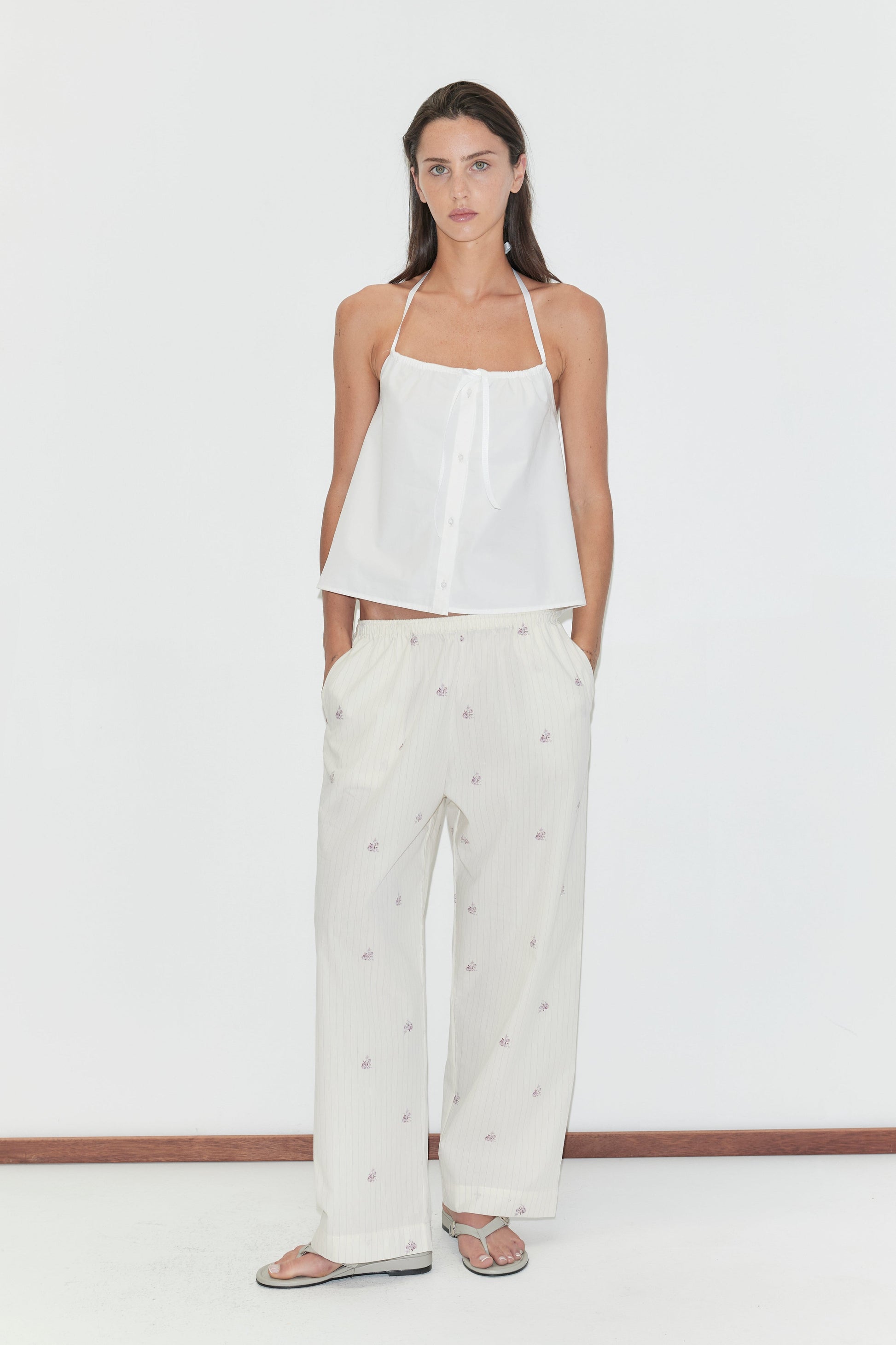 A woman stands against a white background, wearing a white halter top and Deiji Studios’ Ease Trouser - Floral Stripe, featuring a subtle floral stripe pattern. She pairs the look with flip-flops, her hands casually in her pockets, and has long, straight dark hair.