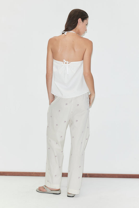 A woman with long dark hair stands with her back to the camera against a plain white background wearing the Simple Tie Top - White by Deiji Studios, a white halter top with an open back and tie fastening, with cream floral trousers.