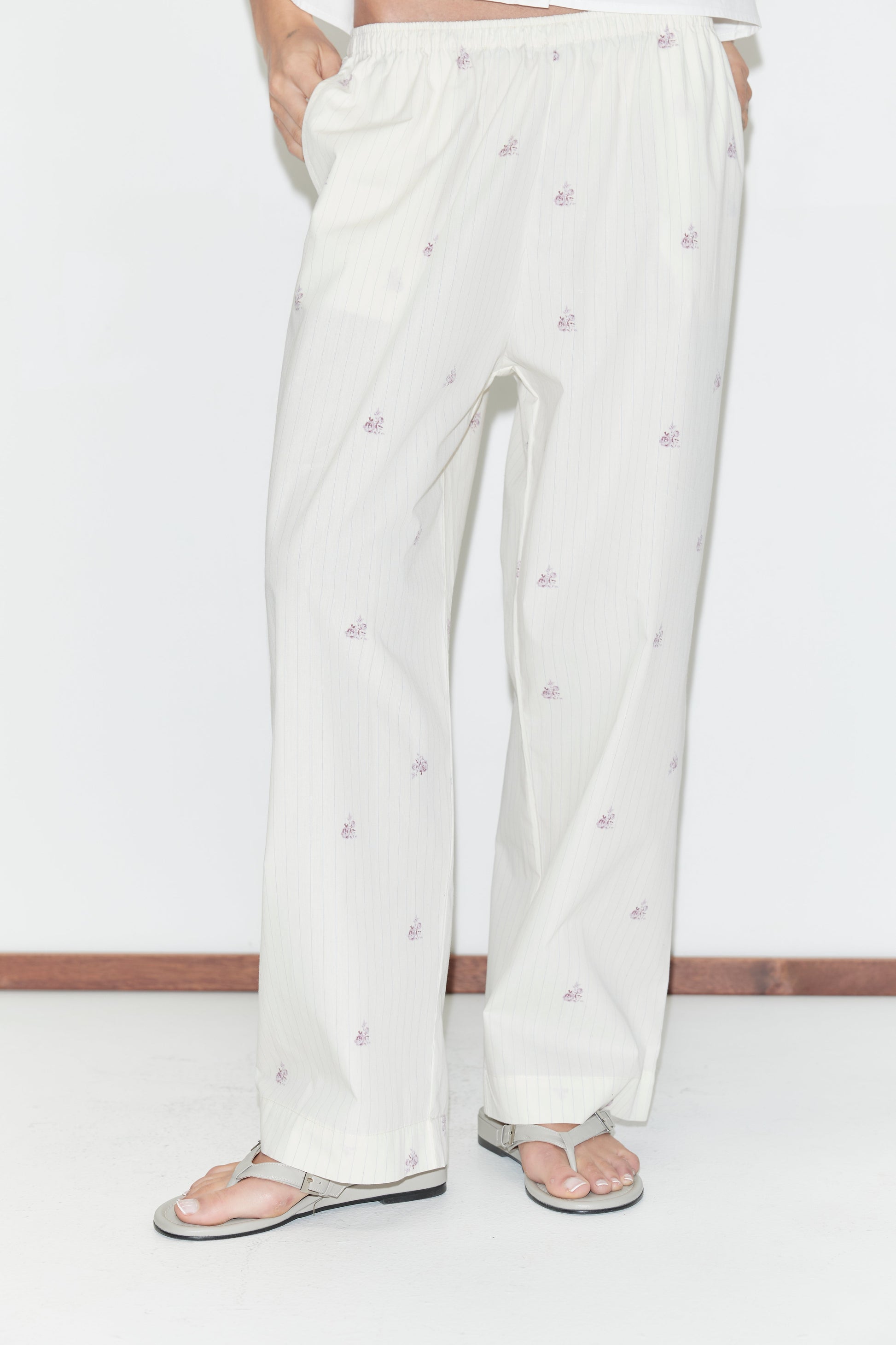 A person is wearing the Ease Trouser - Floral Stripe from Deiji Studios, featuring loose white pajama pants with a subtle pink floral stripe pattern, standing on a white floor with sandals; their upper body is partially out of frame.